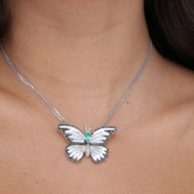 Close-up of a model wearing an 18-karat gold butterfly pendant with white enamel wings, accented by black diamonds, and featuring a 2.55-carat Colombian emerald at the center, suspended on a fine double chain.